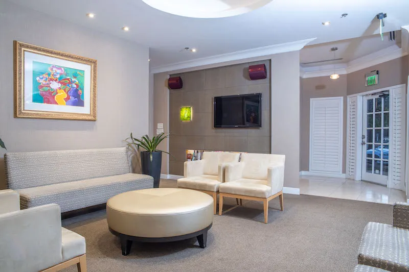 A modern, high-end dental office waiting area with cream-colored sofas, a round ottoman, and framed artwork on the wall. The lighting is warm and professional