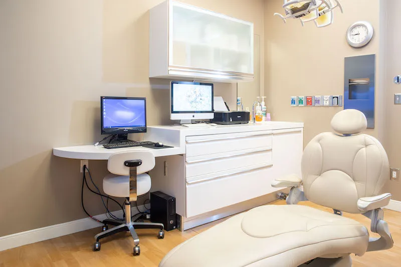 A high-tech dental operatory with a white patient chair, dual computer monitors, and sleek cabinetry
