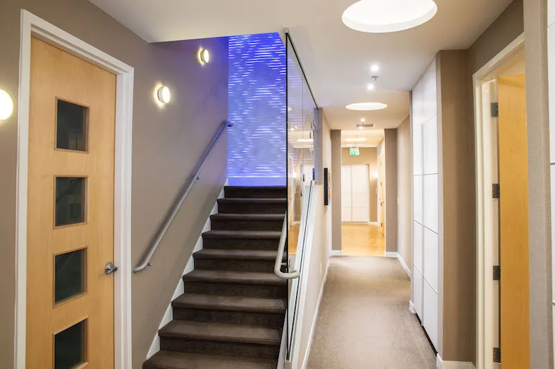 A modern office hallway featuring a dark wood staircase with a vibrant blue backlit wall panel