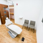 A clinical operatory room featuring a modern patient chair, hardwood floors, and a wall-mounted monitor