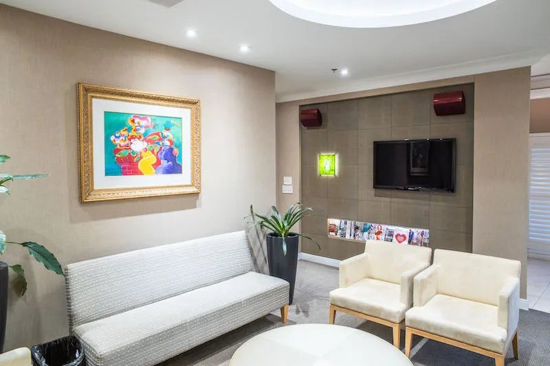 A patient waiting lounge with white leather seating, a wall-mounted TV, and framed colorful art