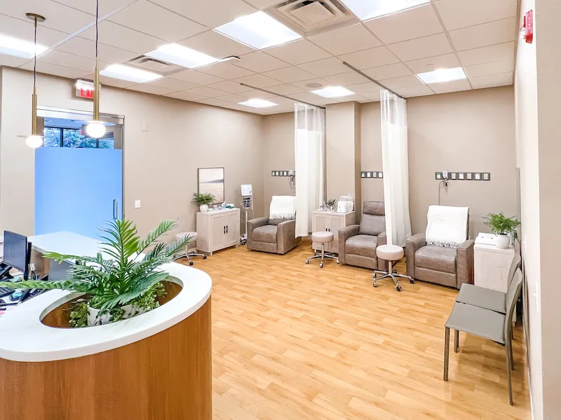 A clean dental waiting room featuring wood-style flooring, a curved white reception desk with a built-in planter, and gray armchairs