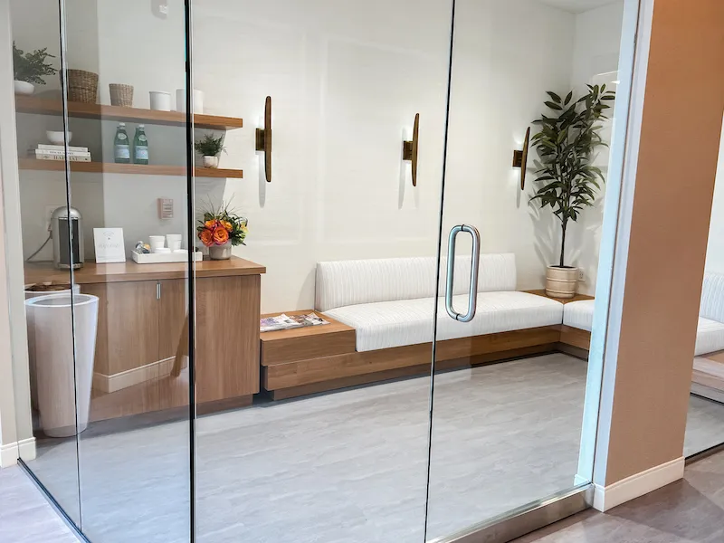View through glass doors into a clinical workspace with wooden cabinetry and a large plant