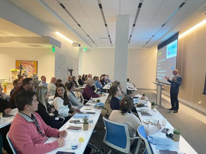 An overhead view of a classroom-style seminar with attendees sitting at desks facing a presenter and a projector screen