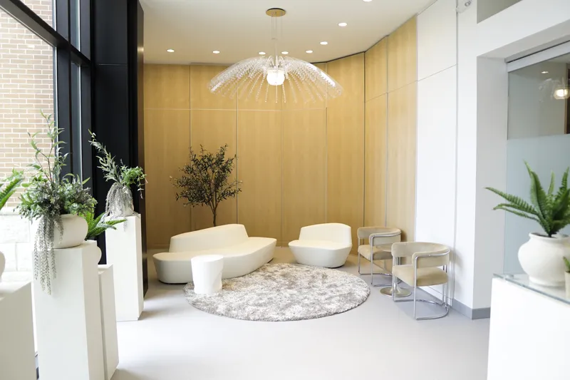 A patient lounge area with neutral-toned walls, decorative plants in white tall planters, and plenty of natural light from floor-to-ceiling windows