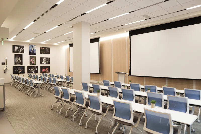 A large conference room or classroom setup with rows of blue chairs facing a large projection screen