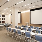 A large conference room or classroom setup with rows of blue chairs facing a large projection screen