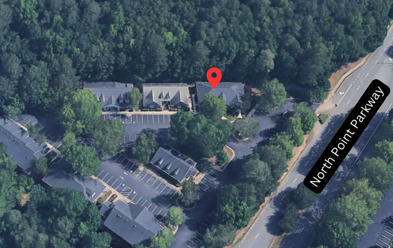 An aerial satellite view of a commercial office park surrounded by trees, with a red pin marking the specific building location