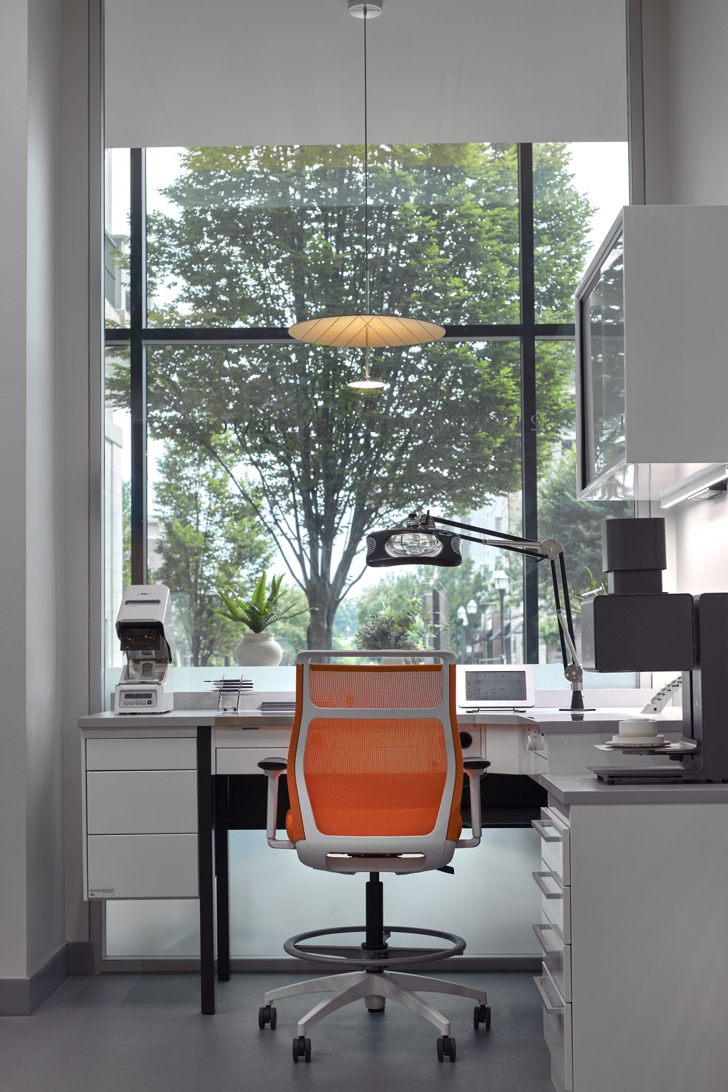 A clean office workstation with a computer, an orange ergonomic chair, and a large window overlooking trees